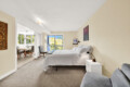 Bedroom with white bedding and beige walls; a nightstand sits beside the bed, with a view into a bright dining area through large windows.