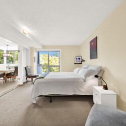 Bedroom with white bedding and beige walls; a nightstand sits beside the bed, with a view into a bright dining area through large windows.
