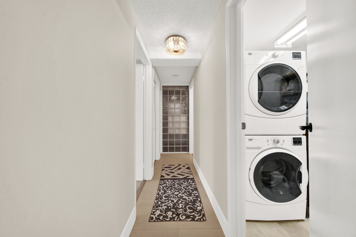 Narrow hallway with a stacked washer/dryer on the right, beige walls, and a patterned rug running down the floor.