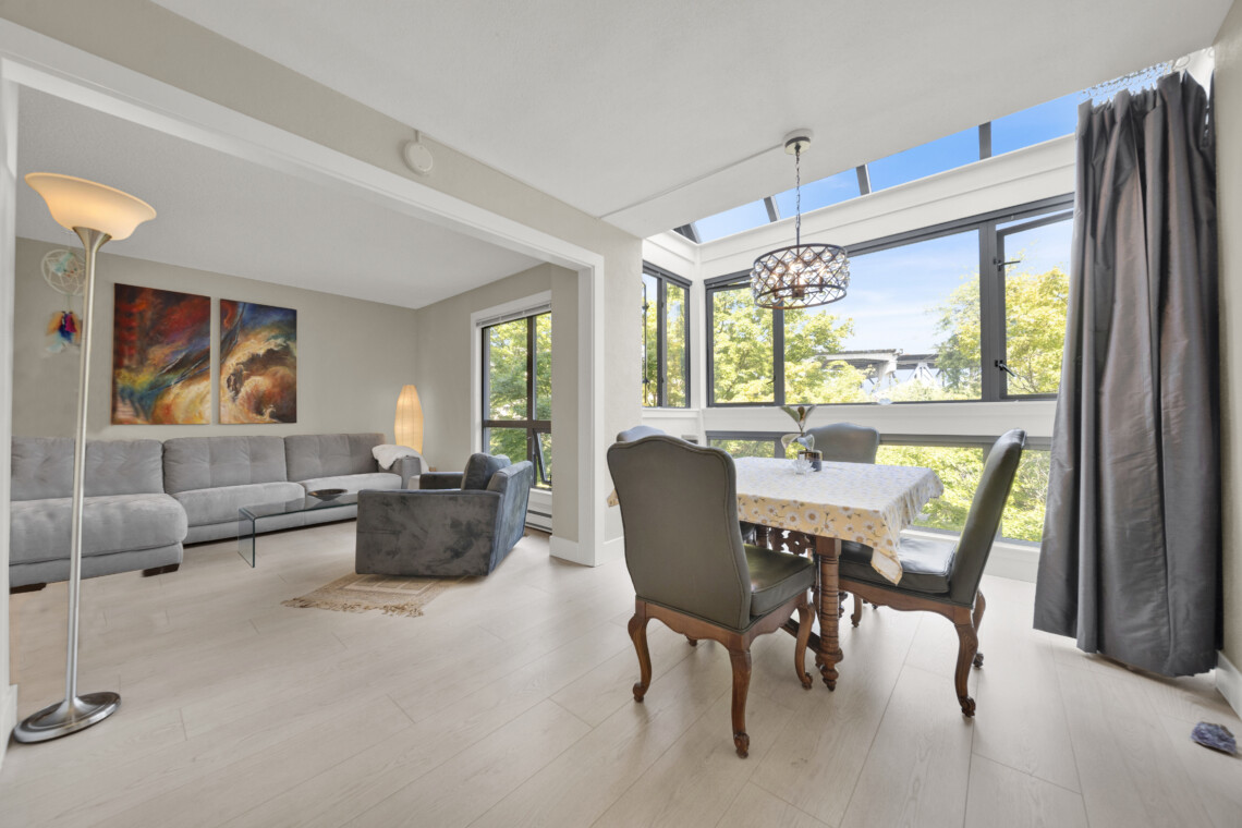 Bright living-dining space with a grey sectional sofa, abstract wall art, and a tall floor lamp near large windows.