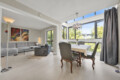 Bright living-dining space with a grey sectional sofa, abstract wall art, and a tall floor lamp near large windows.