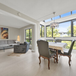 Bright living-dining space with a grey sectional sofa, abstract wall art, and a tall floor lamp near large windows.