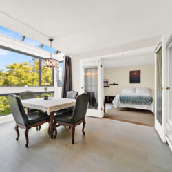 Sunlit dining area with a wooden table and six dark chairs beside large windows overlooking trees; doorway leads to a bedroom beyond.