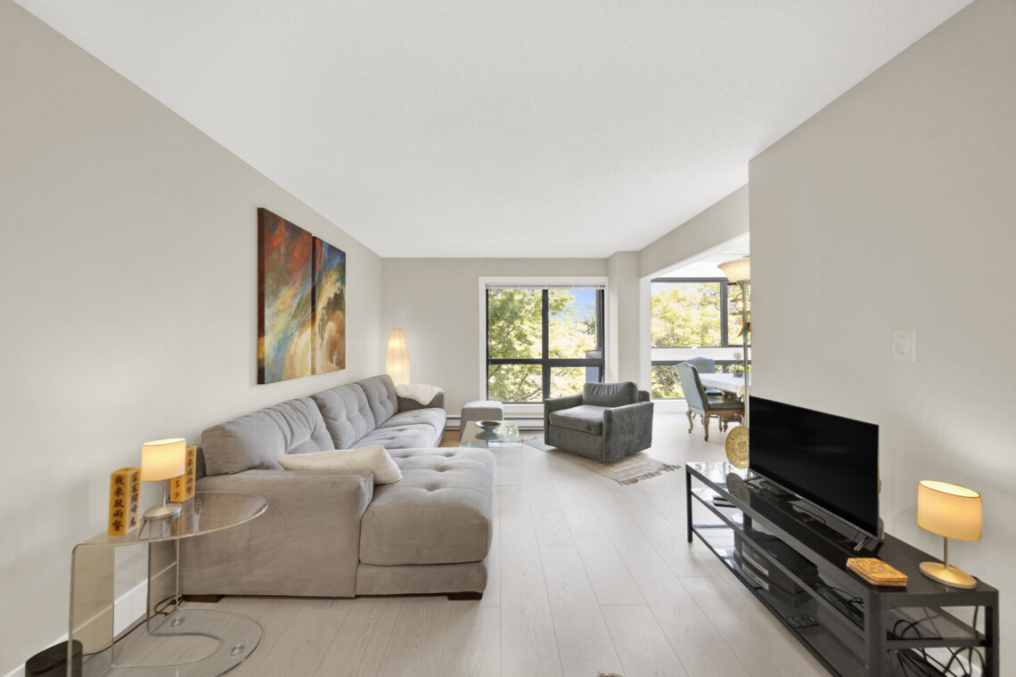 Bright living room with a light gray sectional, glass coffee tables, abstract wall art, and large windows showing greenery.