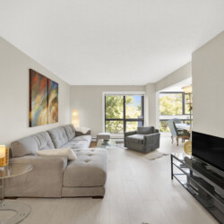 Bright living room with a light gray sectional, glass coffee tables, abstract wall art, and large windows showing greenery.