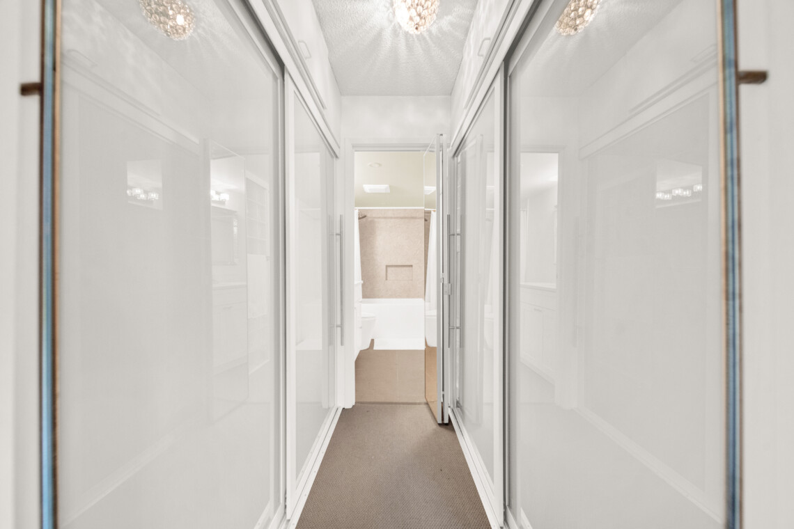 Narrow hallway with glass-front cabinets on both sides, reflecting light from ceiling chandeliers and leading to a lit room at the end.