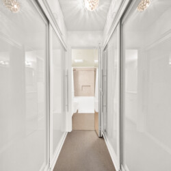 Narrow hallway with glass-front cabinets on both sides, reflecting light from ceiling chandeliers and leading to a lit room at the end.