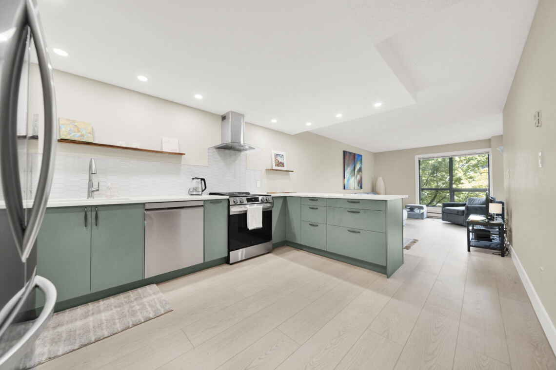 Bright modern kitchen with green lower cabinets, stainless steel appliances, and white tile backsplash; open to a living area with a large window.