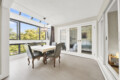 Bright dining room with large windows and skylights, letting in lots of natural light; a table with a yellow floral tablecloth and upholstered chairs around it.