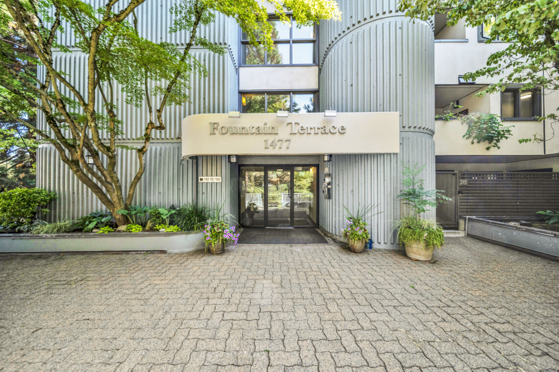 Entrance to a modern residential building with a curved corrugated metal facade, a beige sign reading “Fountain Terrace 1477,” and glass doors framed by greenery and potted plants.