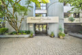 Entrance to a modern residential building with a curved corrugated metal facade, a beige sign reading “Fountain Terrace 1477,” and glass doors framed by greenery and potted plants.