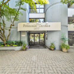 Entrance to a modern residential building with a curved corrugated metal facade, a beige sign reading “Fountain Terrace 1477,” and glass doors framed by greenery and potted plants.