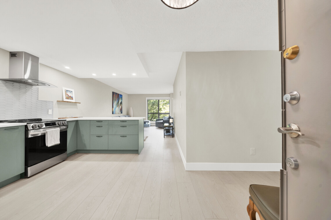 Modern kitchen with mint-green cabinets, stainless steel stove and range hood, and a white tile backsplash; living room visible in the background.
