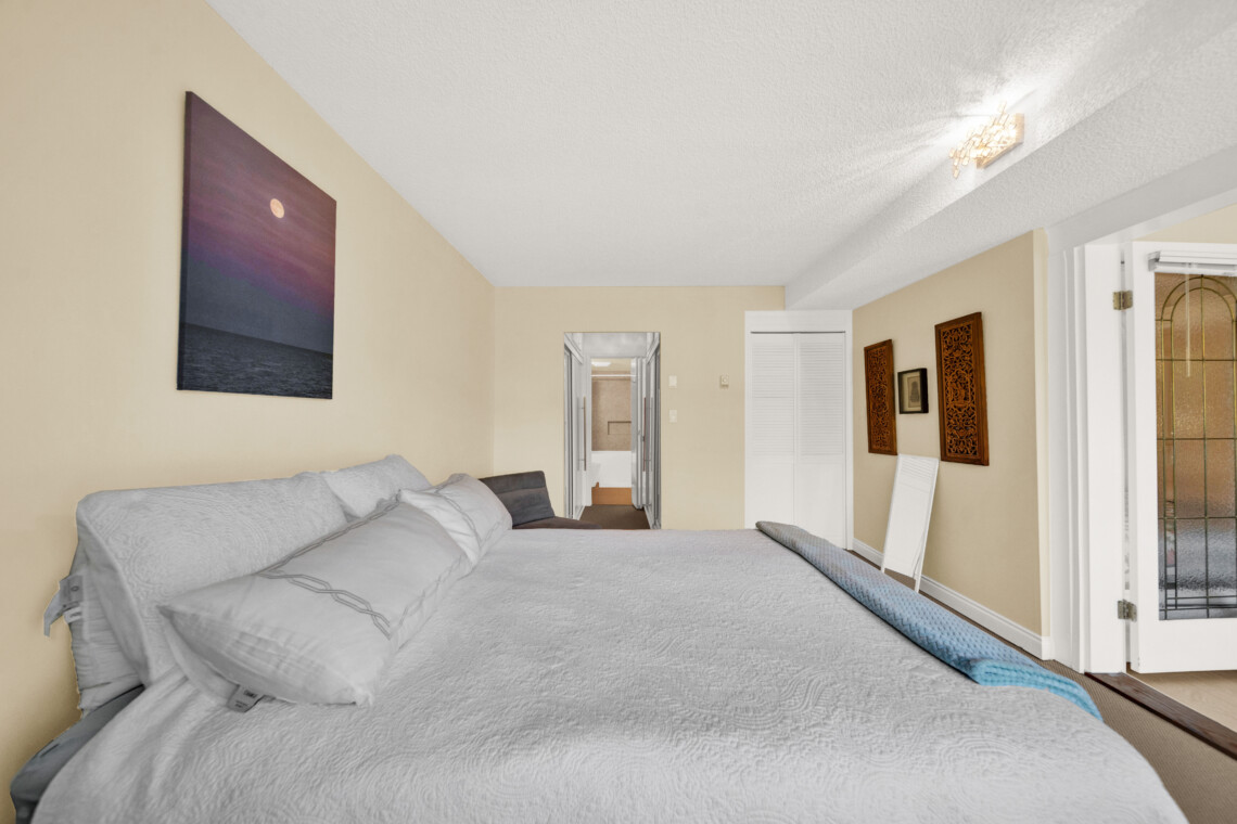 Bedside view of a beige-walled bedroom with white bedding, open doorway to a hallway, and decorative wall panels on the right; balcony door visible at far right.