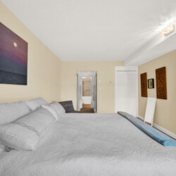 Bedside view of a beige-walled bedroom with white bedding, open doorway to a hallway, and decorative wall panels on the right; balcony door visible at far right.
