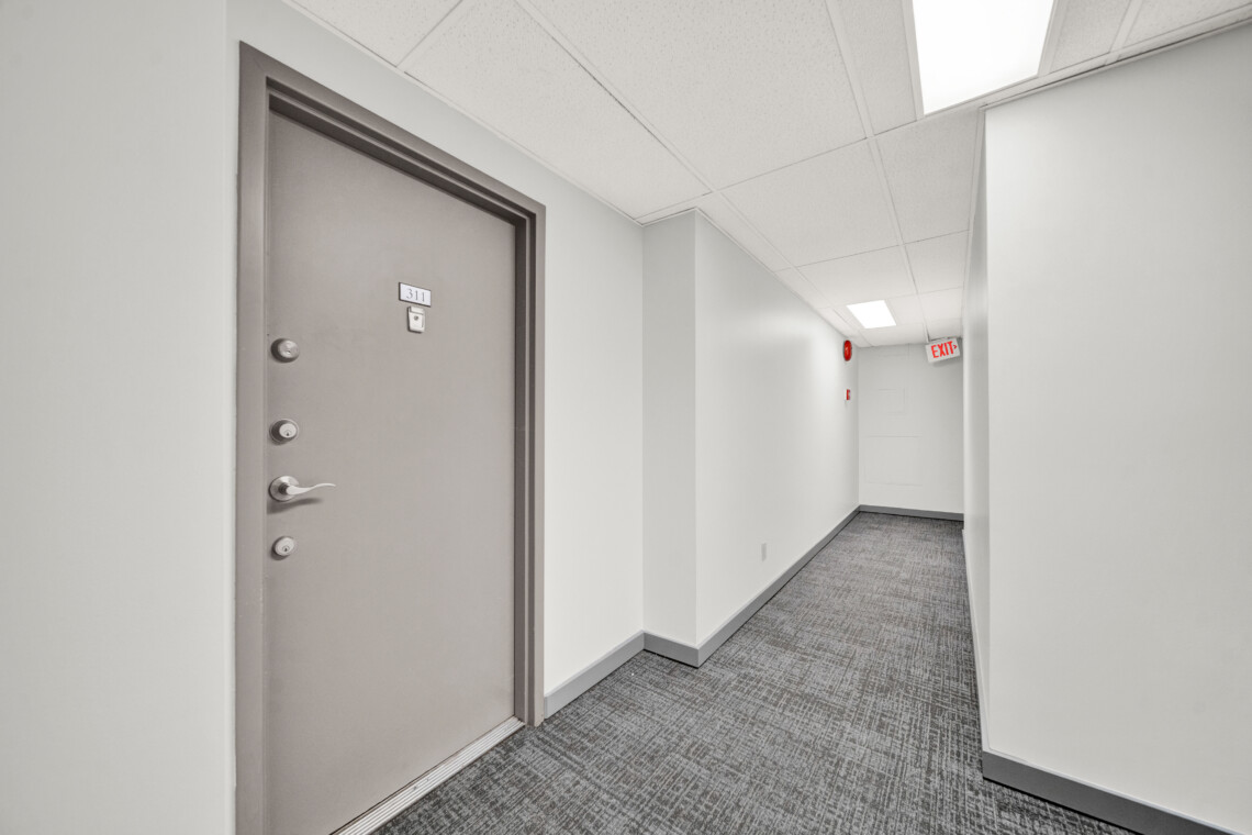 Grey hallway with a closed door labeled 311 on the left; white walls, carpeted floor, and overhead fluorescent lights; an EXIT sign visible down the hall in the distance