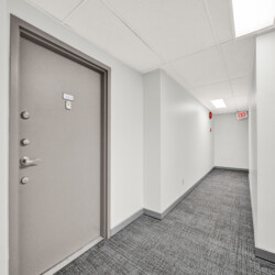 Grey hallway with a closed door labeled 311 on the left; white walls, carpeted floor, and overhead fluorescent lights; an EXIT sign visible down the hall in the distance