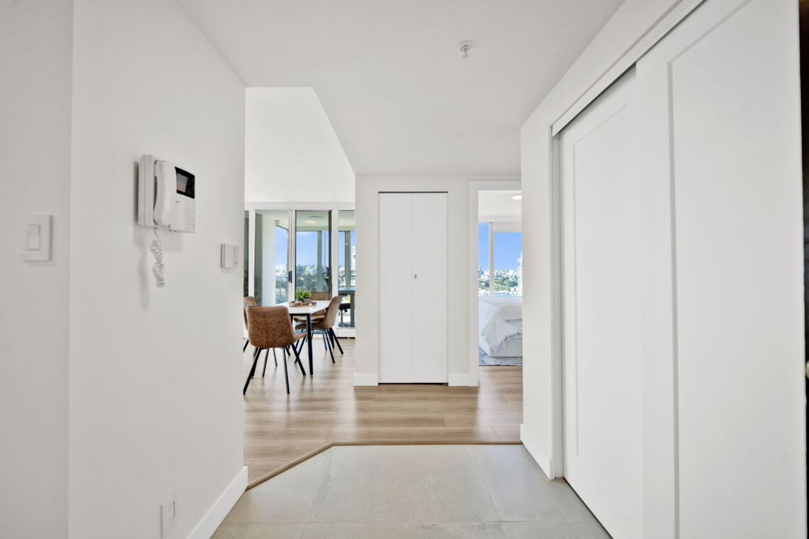 Bright white hallway leading to an open dining area with a wooden table and tan chairs, large glass doors, and city views outside.