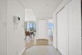 Bright white hallway leading to an open dining area with a wooden table and tan chairs, large glass doors, and city views outside.