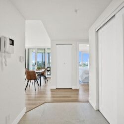 Bright white hallway leading to an open dining area with a wooden table and tan chairs, large glass doors, and city views outside.