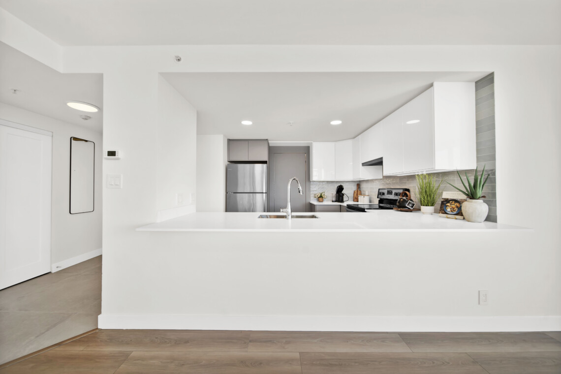 Bright white modern kitchen with an island, stainless steel fridge, and white cabinets; plants on the counter.