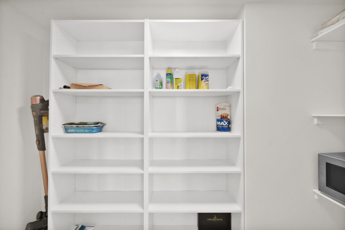 Open white shelving unit in a utility room with cleaning supplies visible on the middle shelf and a vacuum on the left side, leaning against the cabinet.
