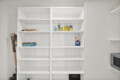 Open white shelving unit in a utility room with cleaning supplies visible on the middle shelf and a vacuum on the left side, leaning against the cabinet.