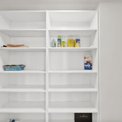 Open white shelving unit in a utility room with cleaning supplies visible on the middle shelf and a vacuum on the left side, leaning against the cabinet.
