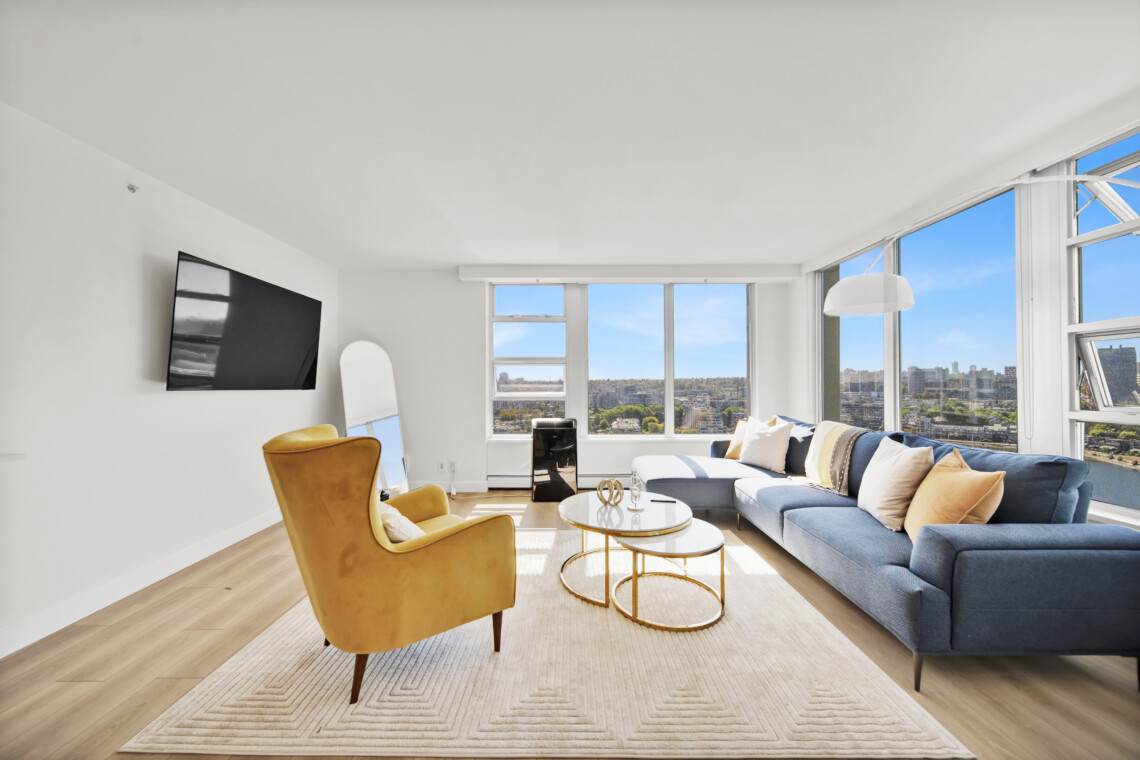 Bright modern living room with large windows overlooking a city, featuring a blue sectional sofa, yellow armchairs, and a round glass coffee table.