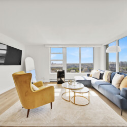 Bright modern living room with large windows overlooking a city, featuring a blue sectional sofa, yellow armchairs, and a round glass coffee table.