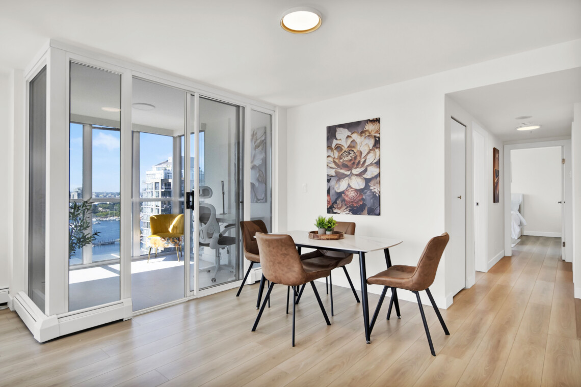 Bright dining area with a glass balcony wall, four tan chairs around a white table, and city view outside.