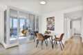 Bright dining area with a glass balcony wall, four tan chairs around a white table, and city view outside.
