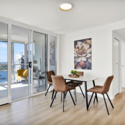 Bright dining area with a glass balcony wall, four tan chairs around a white table, and city view outside.