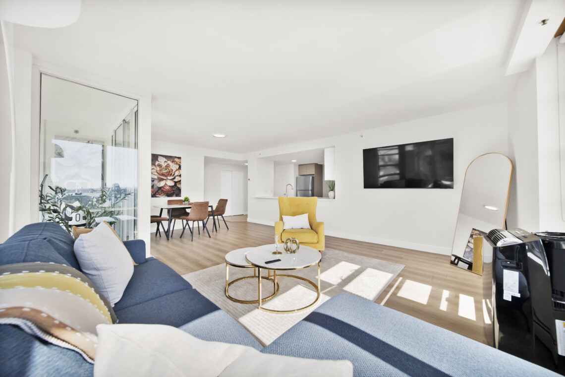 Bright open-concept living area with a blue sofa, yellow armchair, round coffee tables, and a wall-mounted TV on white walls.