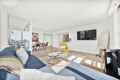 Bright open-concept living area with a blue sofa, yellow armchair, round coffee tables, and a wall-mounted TV on white walls.