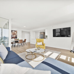 Bright open-concept living area with a blue sofa, yellow armchair, round coffee tables, and a wall-mounted TV on white walls.
