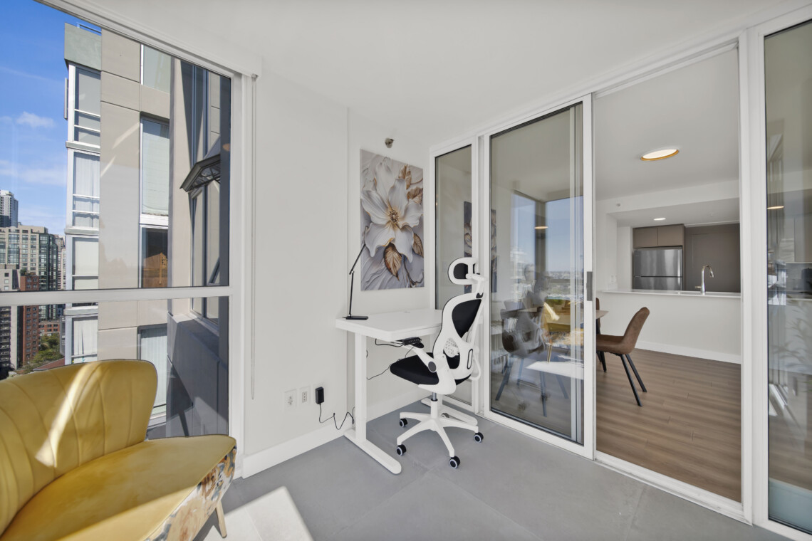 Bright balcony home office with a white desk and ergonomic chair, flower artwork on the wall, and large glass doors opening to a modern kitchen and city views outside.
