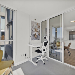 Bright balcony home office with a white desk and ergonomic chair, flower artwork on the wall, and large glass doors opening to a modern kitchen and city views outside.