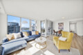 Bright open-plan living room with blue sectional, yellow chairs, and a view of a cityscape through large windows