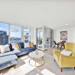 Bright open-plan living room with blue sectional, yellow chairs, and a view of a cityscape through large windows