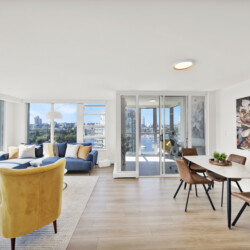 Bright, open living room with large windows showing a city skyline, blue sofa and mustard chairs.