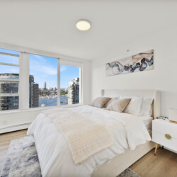 Bright modern bedroom with a large window view of a cityscape and water, white bed and beige throw pillows.