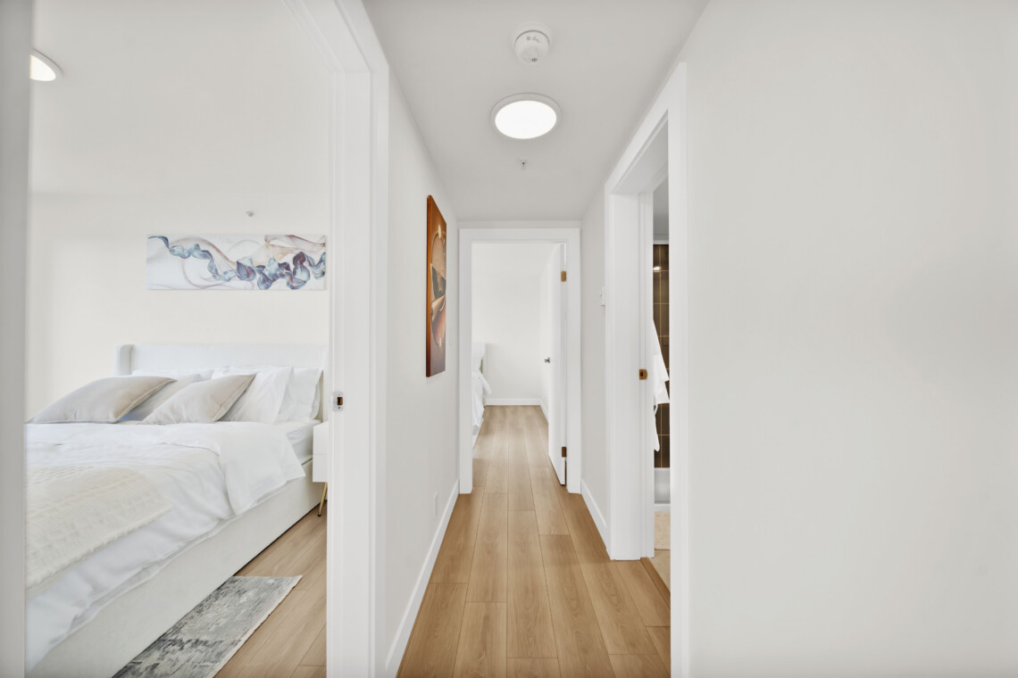 Bright white hallway with open bedroom doors, a bed visible in the left room and light wood flooring.