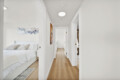 Bright white hallway with open bedroom doors, a bed visible in the left room and light wood flooring.
