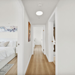 Bright white hallway with open bedroom doors, a bed visible in the left room and light wood flooring.