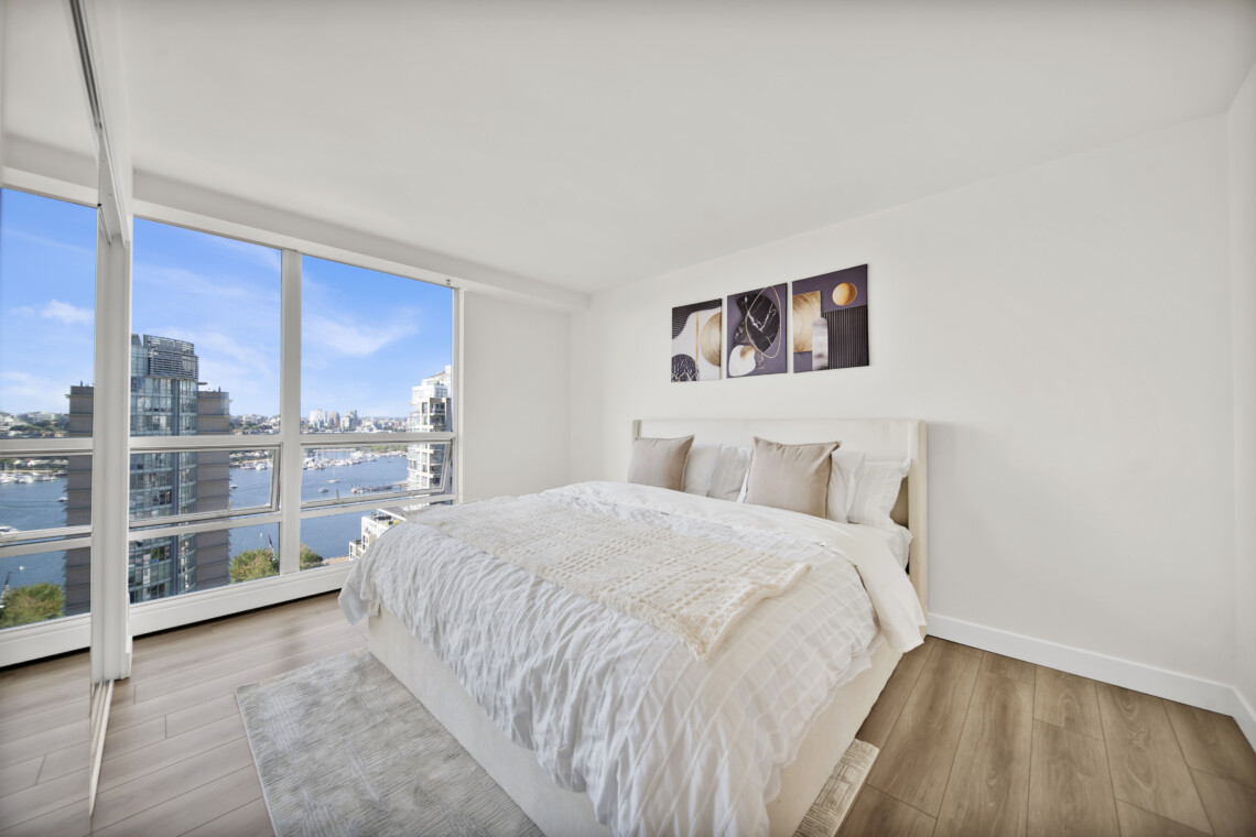 Bright bedroom with a floor-to-ceiling window view of a city harbor and blue sky, a white bed with beige pillows, and abstract triptych wall art.