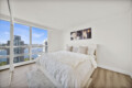 Bright bedroom with a floor-to-ceiling window view of a city harbor and blue sky, a white bed with beige pillows, and abstract triptych wall art.