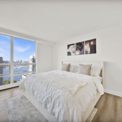 Bright bedroom with a floor-to-ceiling window view of a city harbor and blue sky, a white bed with beige pillows, and abstract triptych wall art.