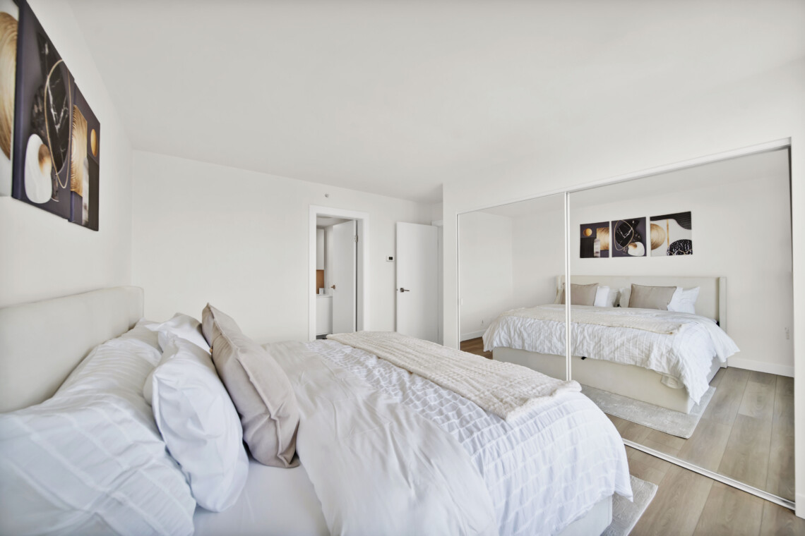Bright modern bedroom with two white-duvet beds, beige pillows, and a mirrored wardrobe reflecting the room.