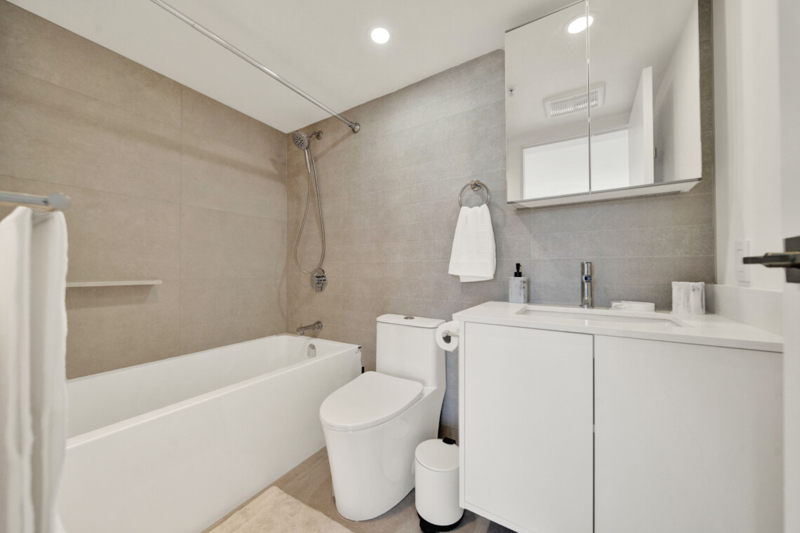 Modern bathroom with a bathtub, handheld shower, beige tiling, and a white vanity with mirrored cabinet above it.